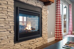 Une cheminée en pierre avec une façade en verre, une poutre en bois au plafond et un rideau à carreaux.