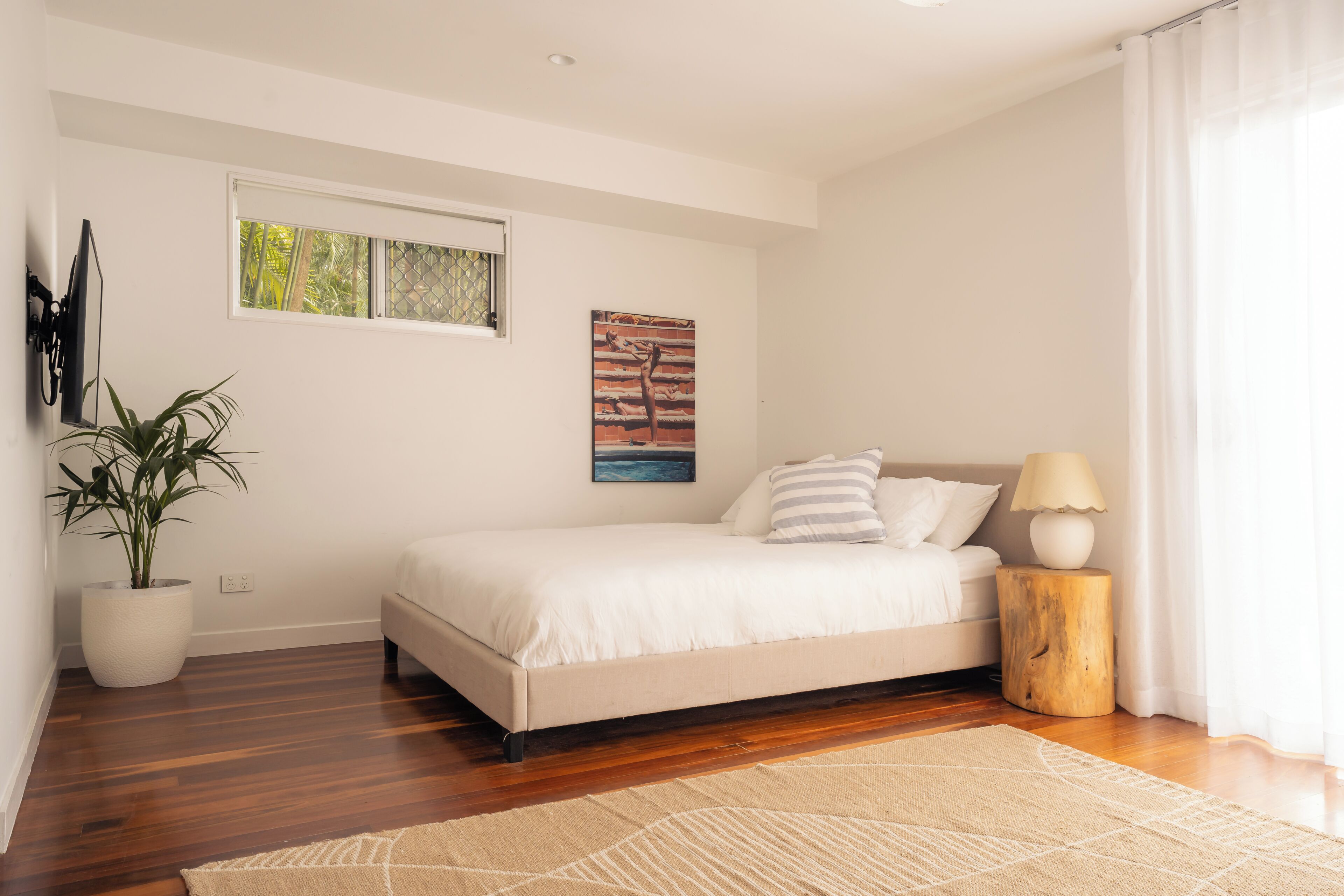 A bedroom with a bed, a potted plant, a painting, and a nightstand with a lamp.