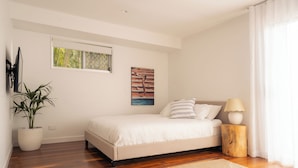 A bedroom with a bed, a potted plant, a painting, and a nightstand with a lamp.