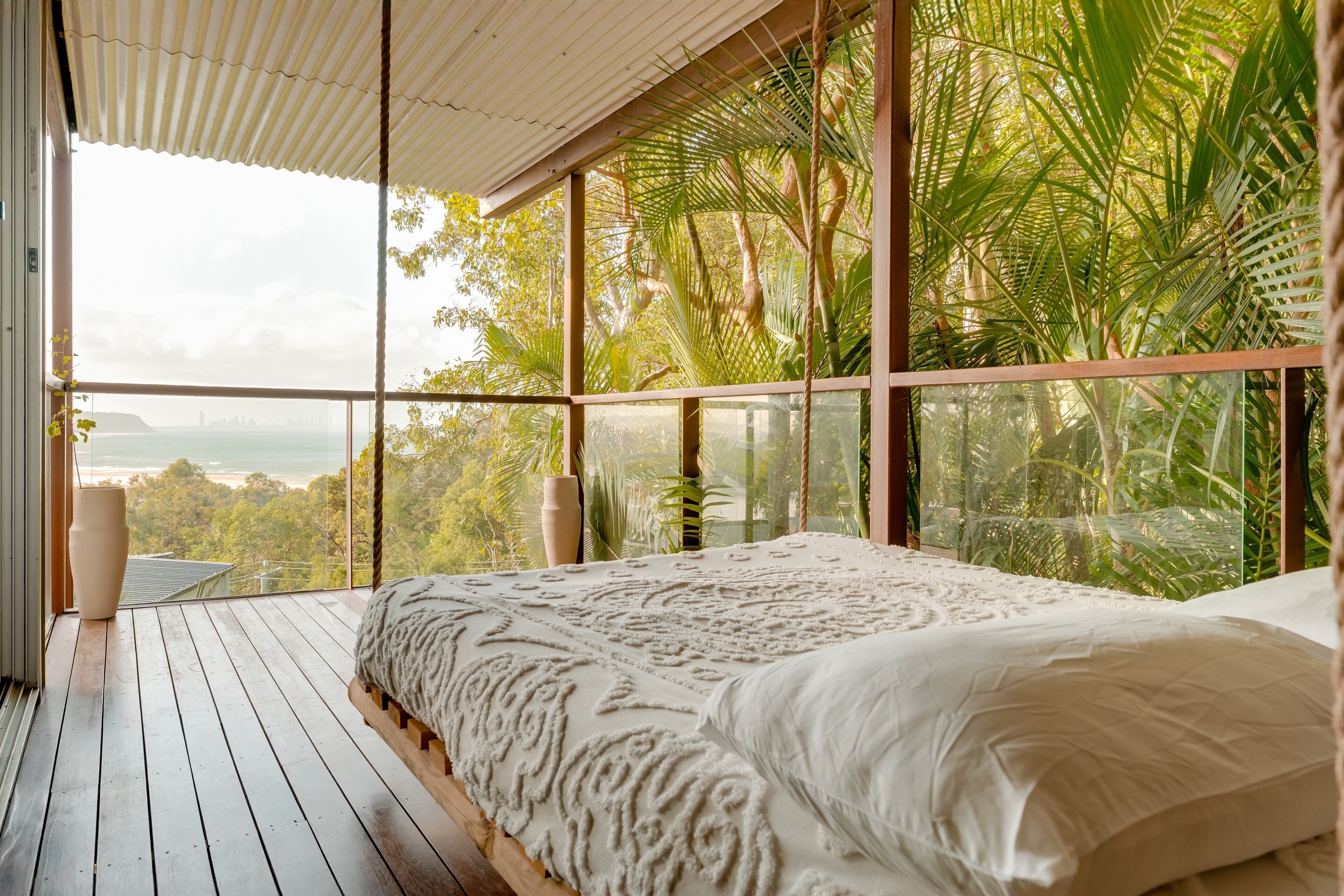 A bedroom with a large bed, wooden flooring, and a view of the outdoors through large windows.