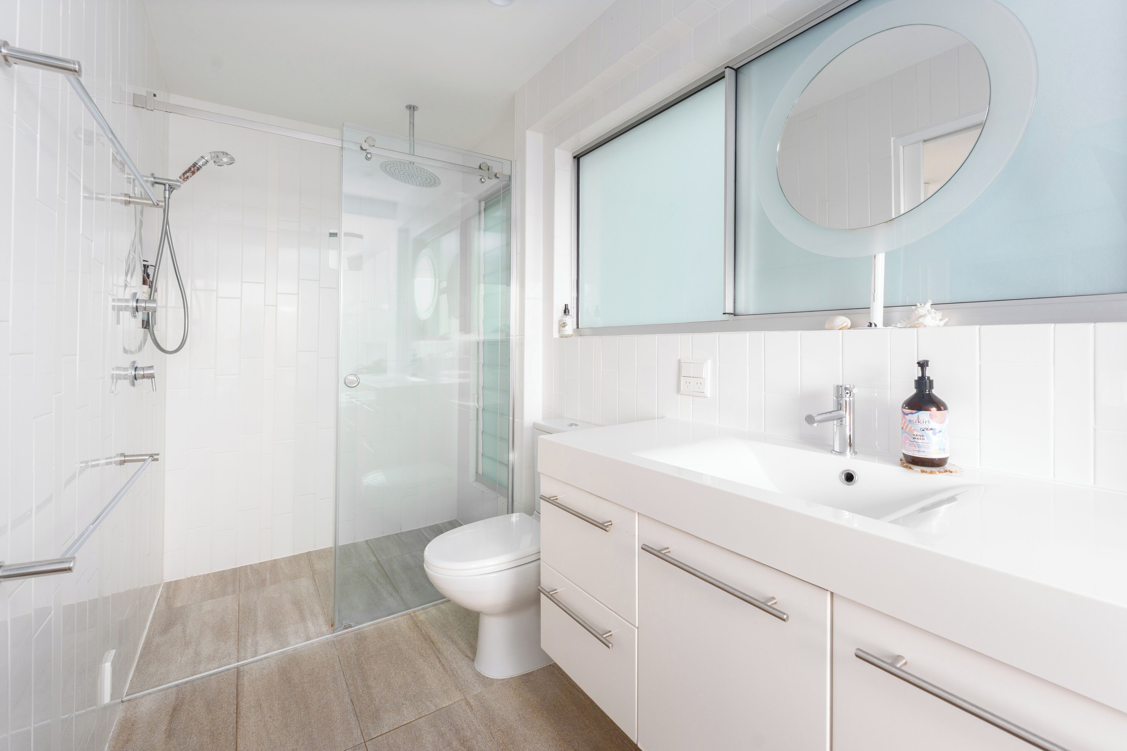A modern bathroom with a glass shower enclosure, a toilet, a sink with a mirror, and a bottle of shampoo on the counter.
