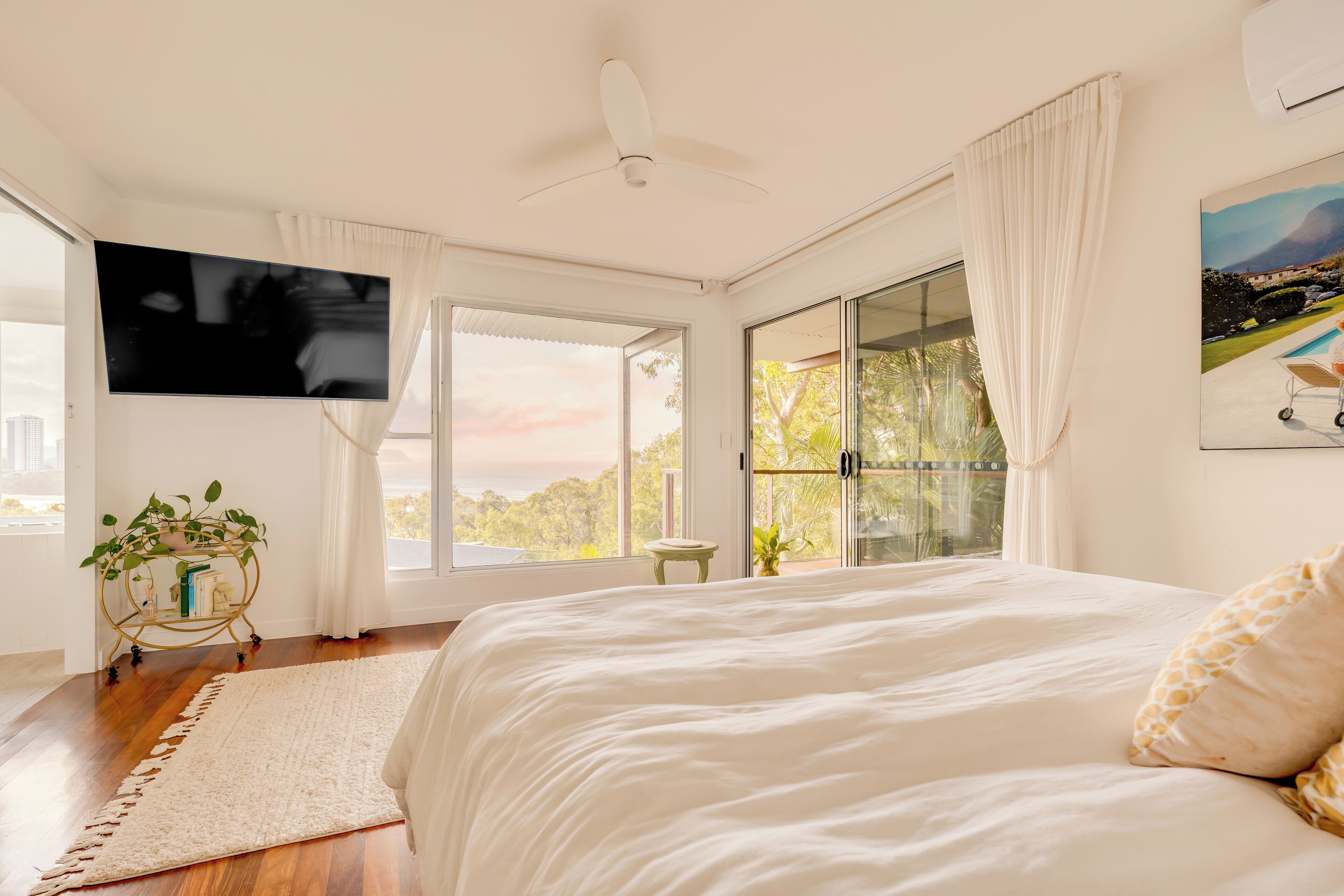 A bedroom with a large bed, a ceiling fan, a TV, and a view of trees through the window.