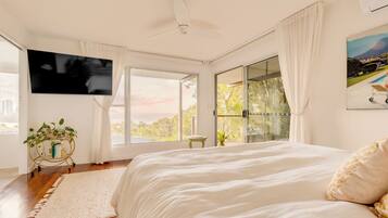 A bedroom with a large bed, a ceiling fan, a TV, and a view of trees through the window.