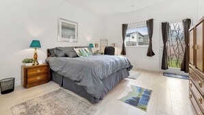 Un dormitorio con cama, mesitas de noche, un escritorio, una silla, una ventana con cortinas y una puerta con felpudo.