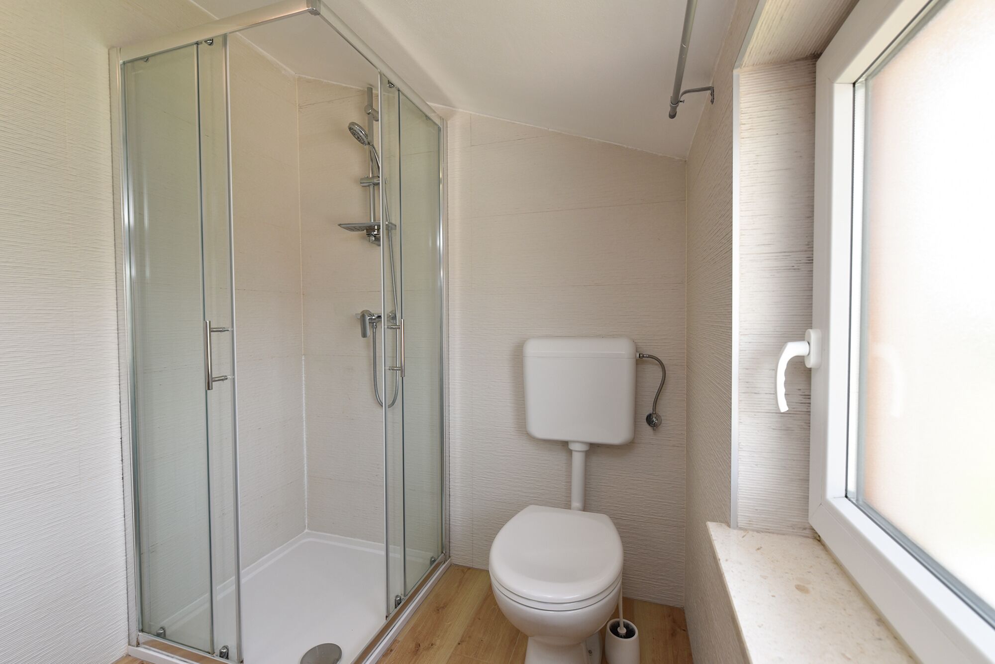 A compact bathroom with a glass shower enclosure, a white toilet, and a window providing natural light.