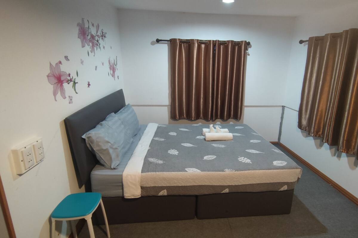 A bedroom with a bed, a small stool, a wall-mounted curtain rod, and a power outlet.