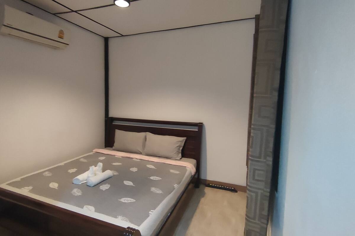 A small, neatly made bed with a leaf-patterned bedspread and pillows, a wooden headboard, a wall-mounted air conditioner, and a window with a patterned curtain.