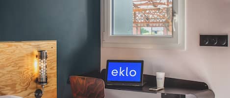 Una habitación de hotel con cama, escritorio con computadora portátil, silla, taza y ventana con vista a un edificio.