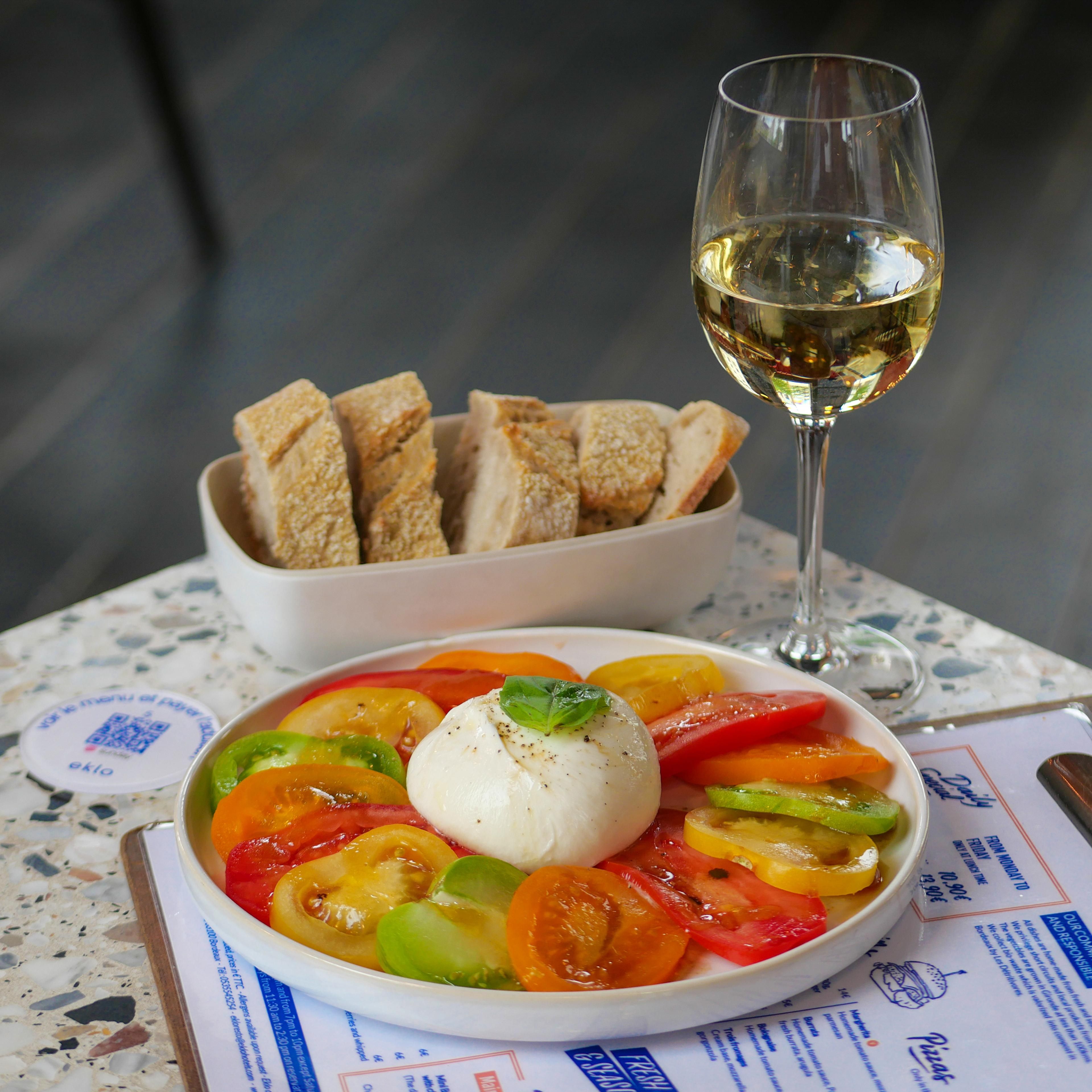 Un plato de ensalada Caprese con tomates, mozzarella y albahaca, una copa de vino blanco y un bol de pan sobre una mesa con un menú.
