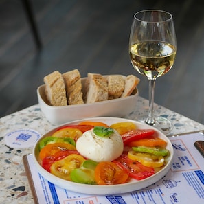 A plate of Caprese salad with tomatoes, mozzarella, and basil, a glass of white wine, and a bowl of bread on a table with a menu. - Eklo Hotels Paris Roissy CDG Airport (Roissy-en-France)