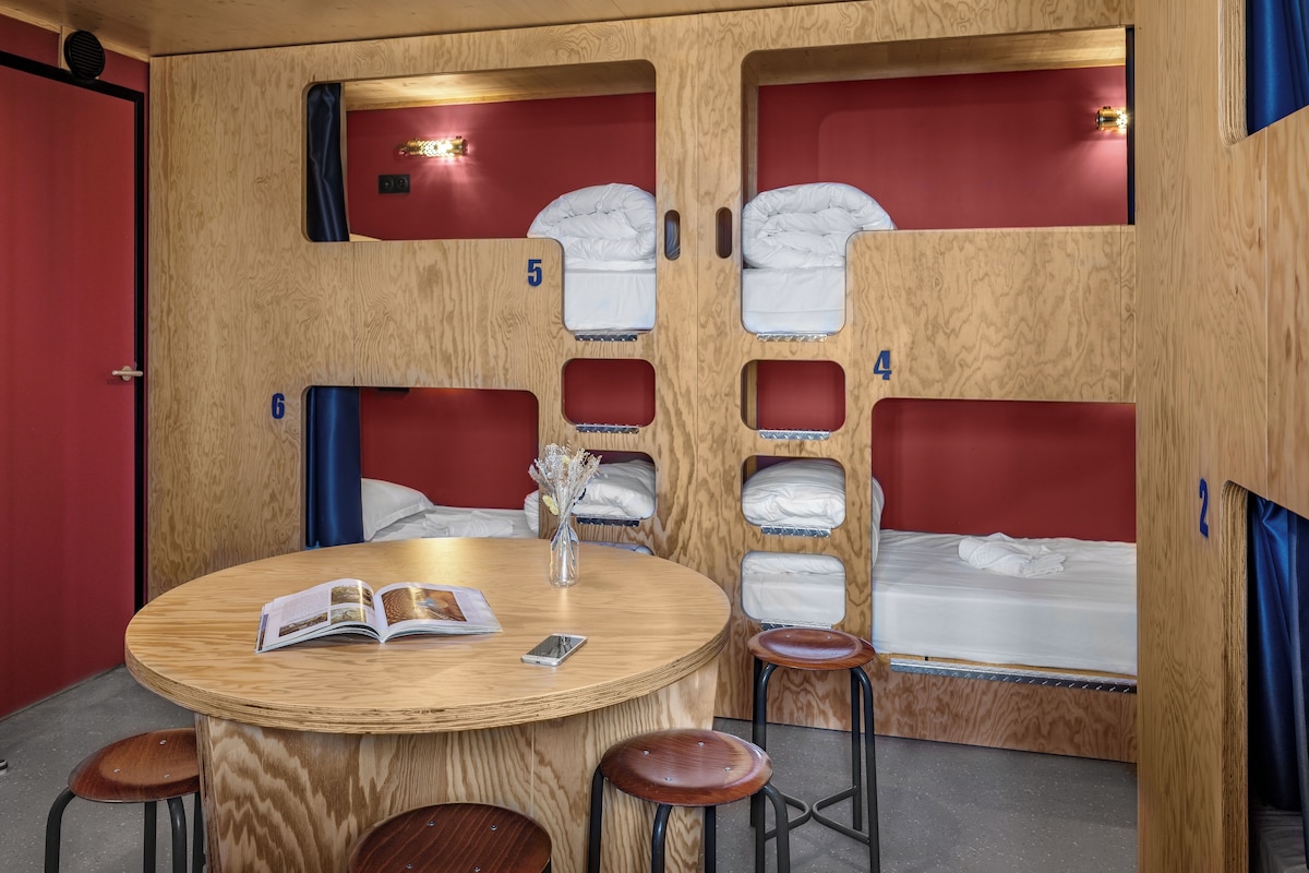 A room with bunk beds numbered 2, 4, 5, and 6, a round wooden table with a book and a vase, and wooden stools.