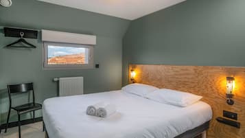 Una cama bien tendida con sábanas blancas, un cabecero de madera, una mesita de noche con una lámpara, una silla, una ventana con vista a un edificio y un radiador.