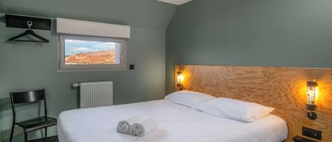 Una cama bien tendida con sábanas blancas, un cabecero de madera, una mesita de noche con una lámpara, una silla, una ventana con vista a un edificio y un radiador.