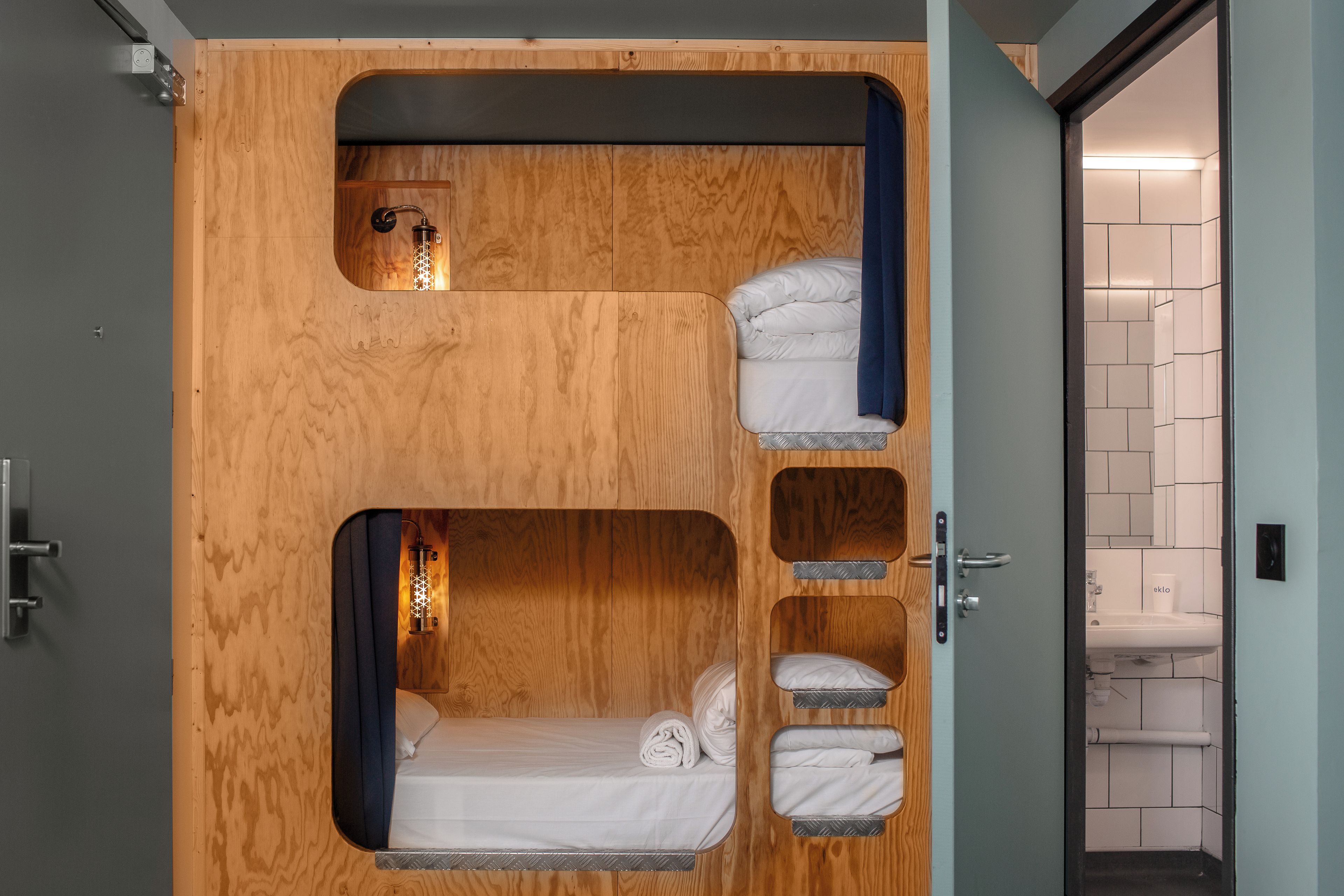 Una habitación pequeña con literas, una ventana discreta y un baño a la vista, accesible a través de una puerta abierta.