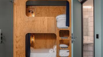 Una habitación pequeña con literas, una ventana discreta y un baño a la vista, accesible a través de una puerta abierta.