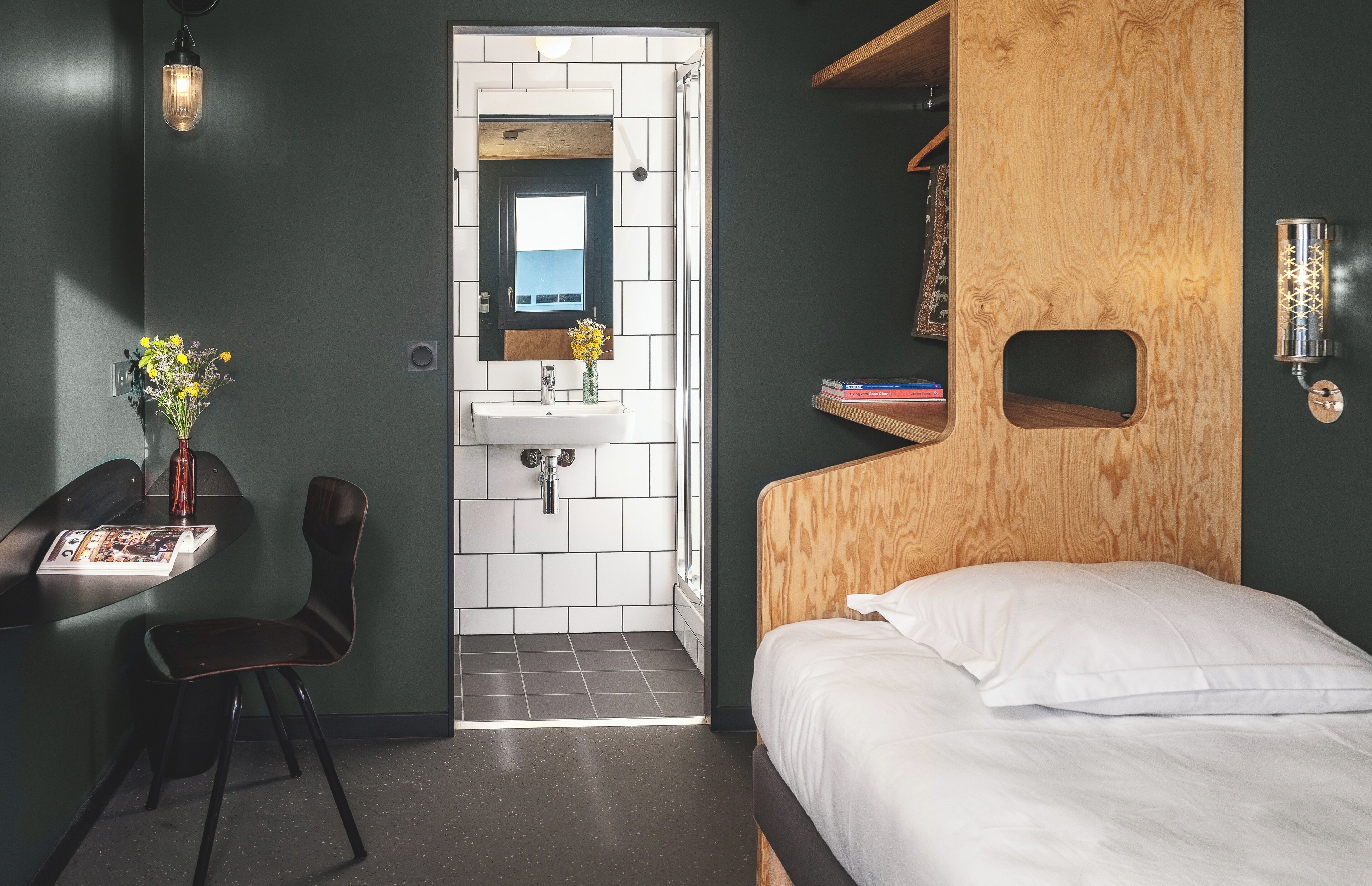 Une chambre d’hôtel de taille modeste, dotée d’une tête de lit en bois, d’un petit lavabo, d’une chaise et d’une table de chevet avec un magazine.