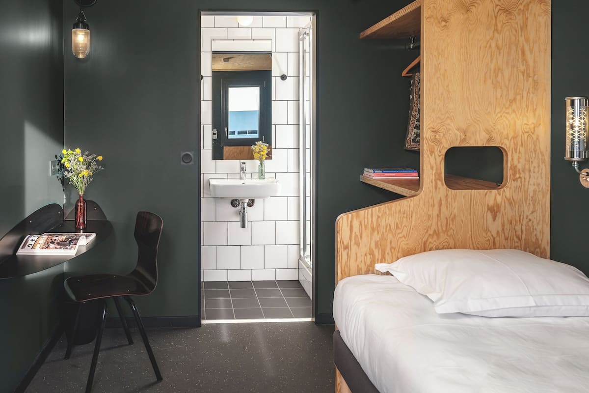 A compact room with a bed, a small desk with a chair, a wall-mounted shelf, a wooden headboard, a white sink, and a mirror.