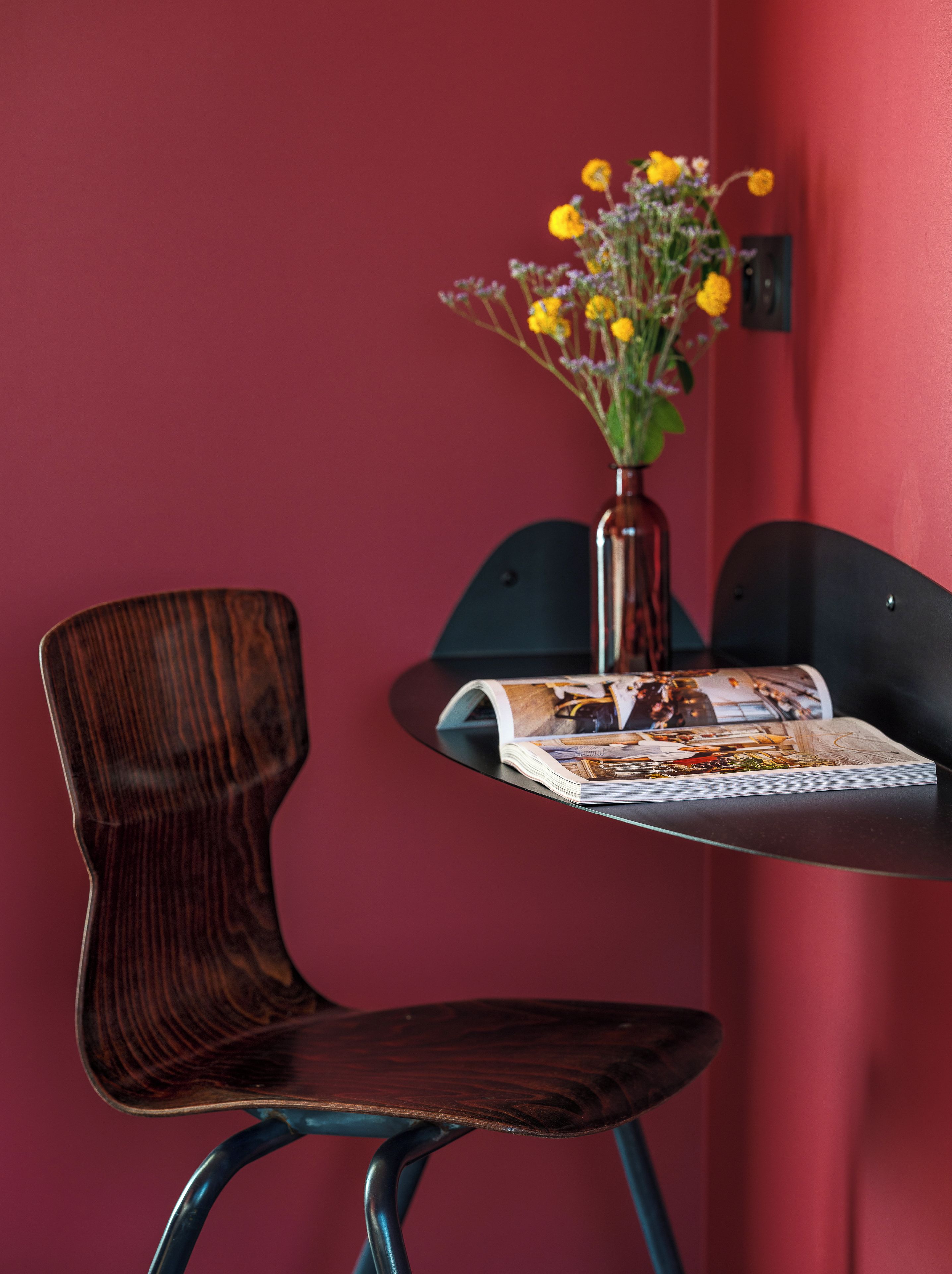 Una silla de madera con respaldo curvo y patas metálicas, una mesita con una revista, y un florero con flores amarillas frente a una pared roja.