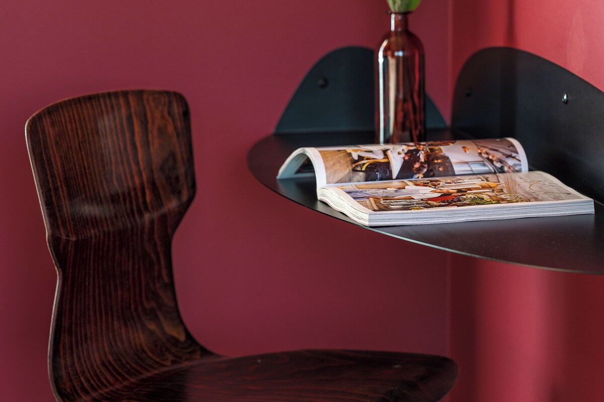 A wooden chair with a curved backrest and metal legs, a small table with a magazine, and a vase with yellow flowers against a red wall.