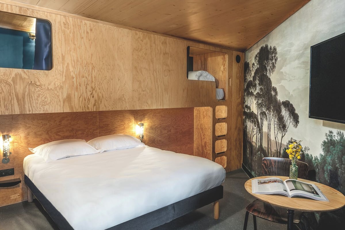 A wooden room with a bed, a small table with a book, and a wall-mounted TV.