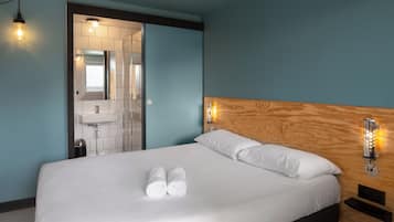 Una habitación de hotel con una cama, cabecera de madera, mesita de noche, lámpara de pared y un baño visible a través de una puerta abierta.