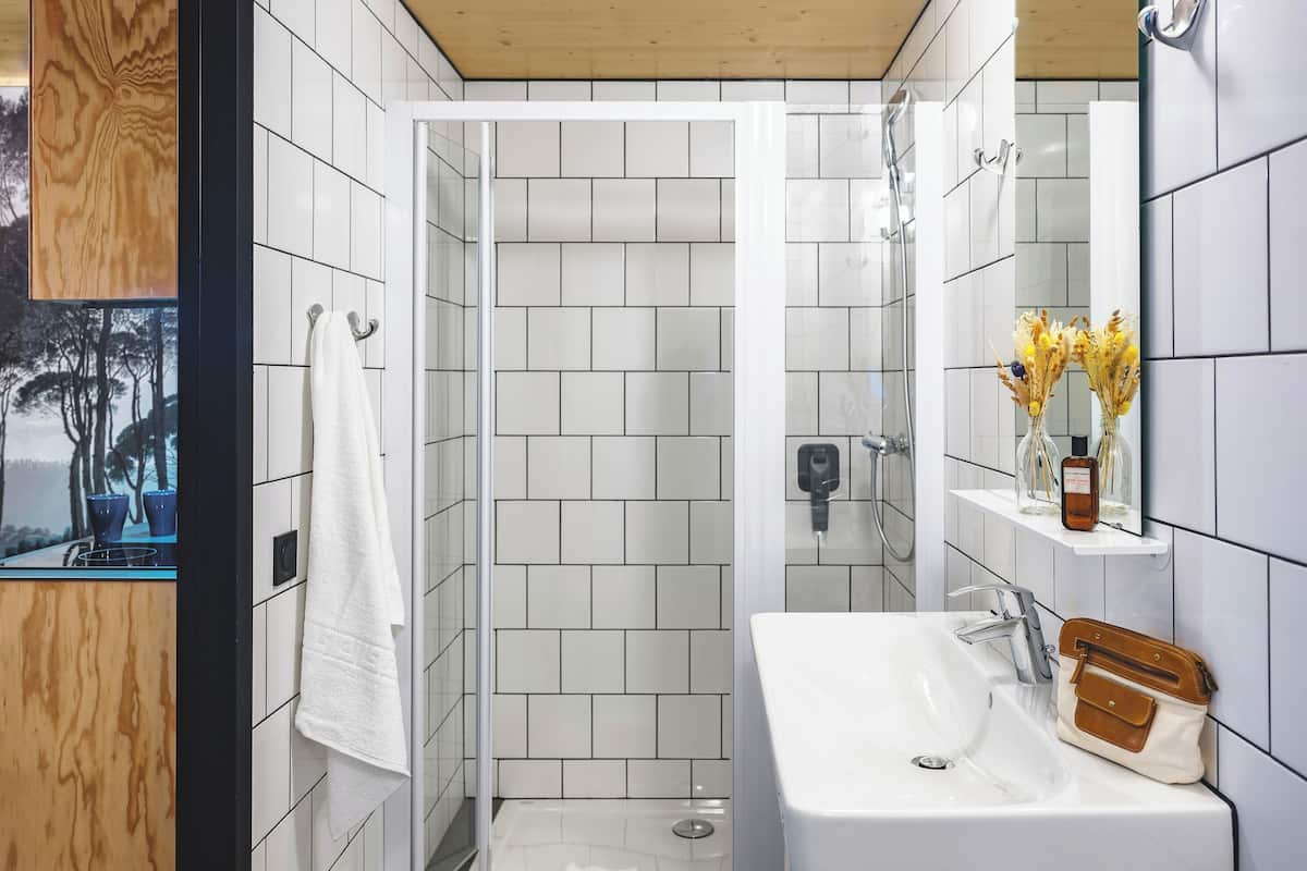 A modern bathroom with white tiles, a wooden vanity, a shower enclosure, and a mirror with a vase of flowers.