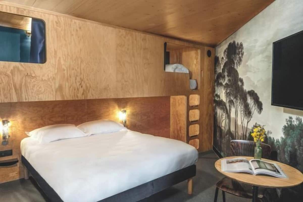 A bedroom with a bed, bedside lamps, a small table with a book, and a wall-mounted shelf.
