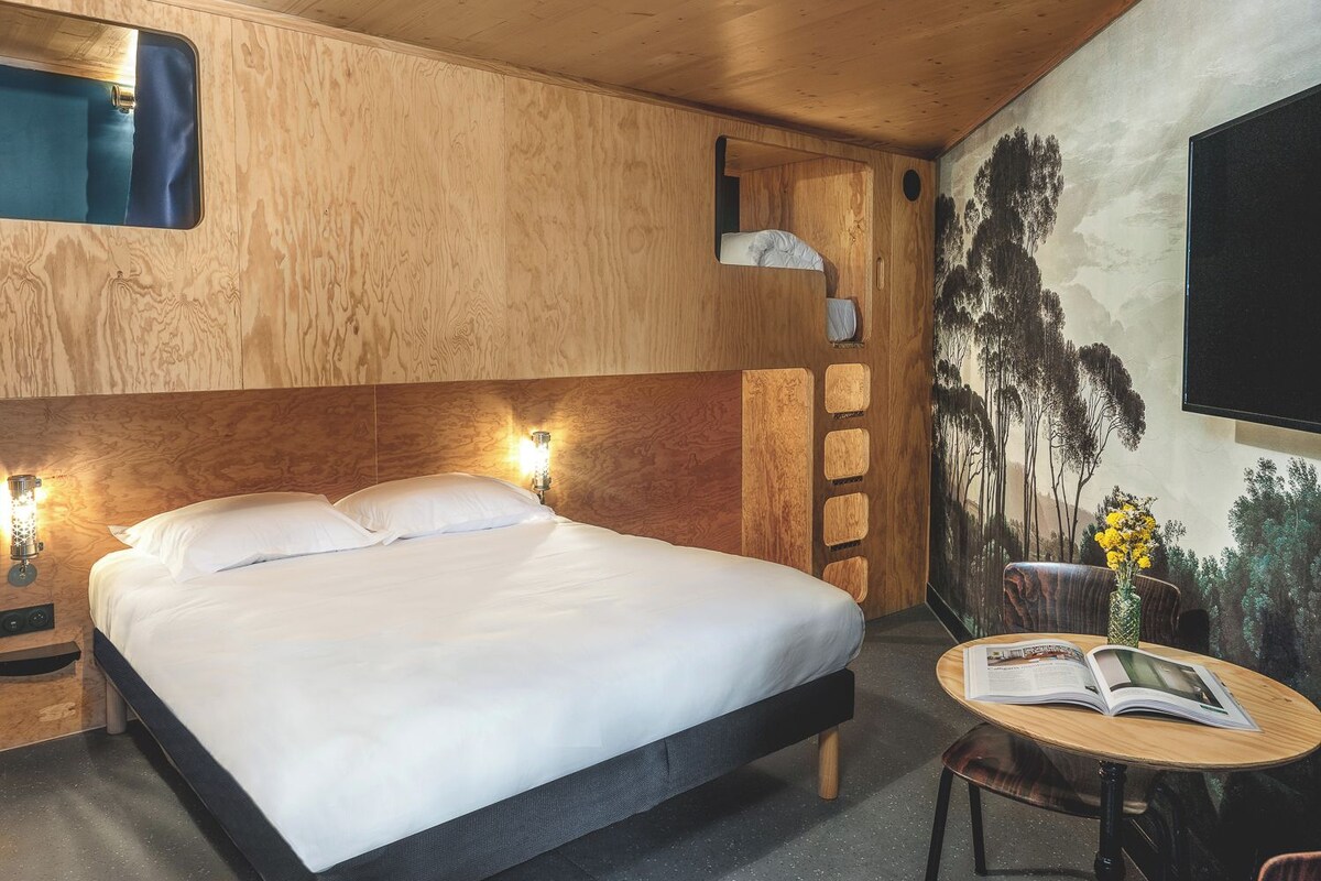 A wooden room with a bed, a small table with a book, and a wall-mounted TV.