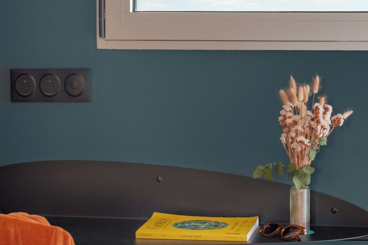 A room with a teal wall, a window with a view of the sky, a black shelf with a book and a vase of dried flowers, and a bed with orange and white bedding.