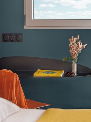 Una habitación con pared verde azulado, ventana con vista al cielo, estante negro con un libro y un jarrón de flores secas, y cama con ropa de cama naranja y blanca.
