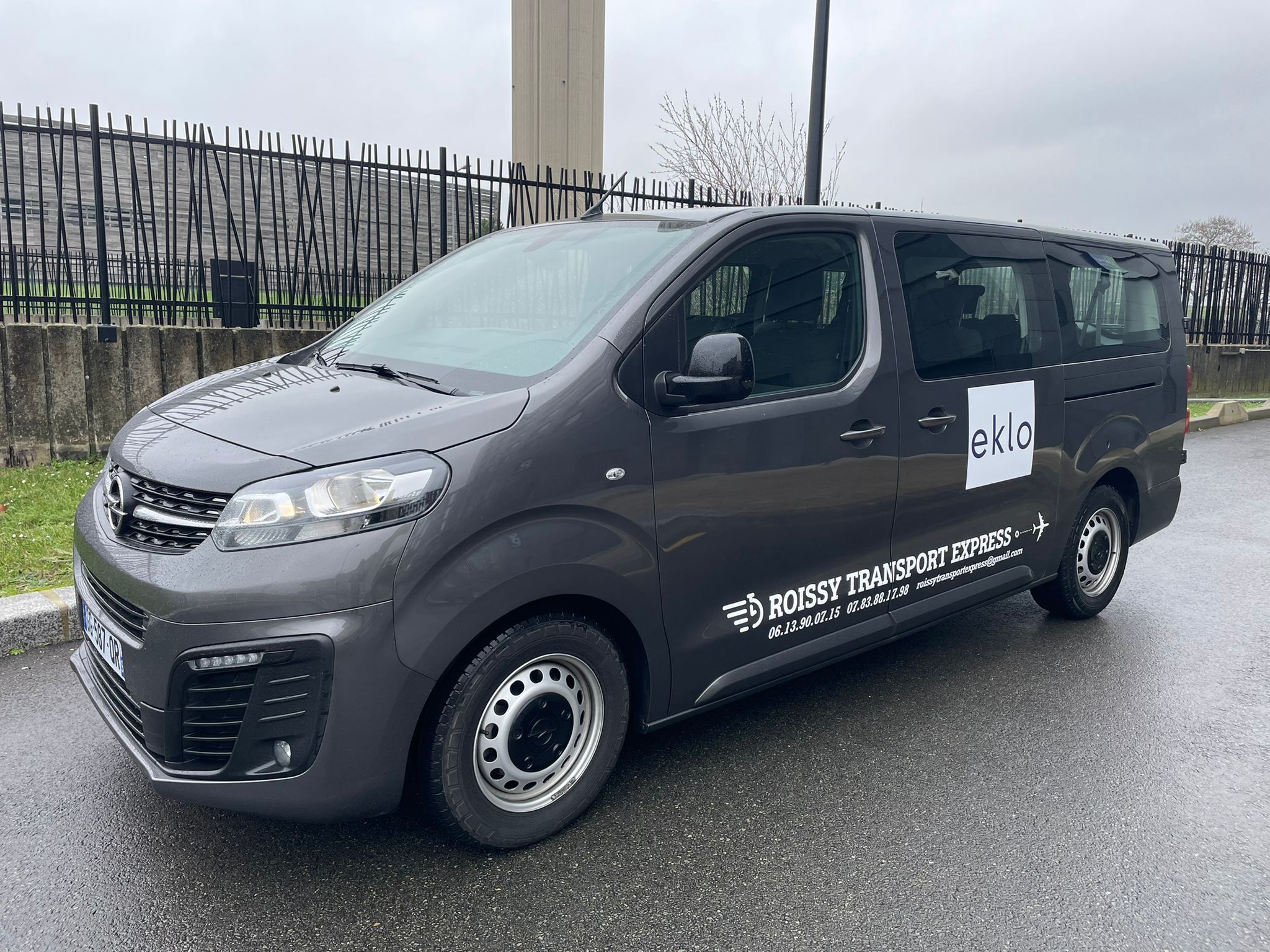 Un fourgon gris portant les inscriptions « ROISSY TRANSPORT EXPRESS » et « eklo » était garé sur une route mouillée.