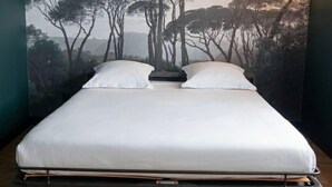 A bed with a white mattress, a headboard, and a pull-out section, set against a wall with a forest landscape mural. - Eklo Hotels Paris Roissy CDG Airport (Roissy-en-France)