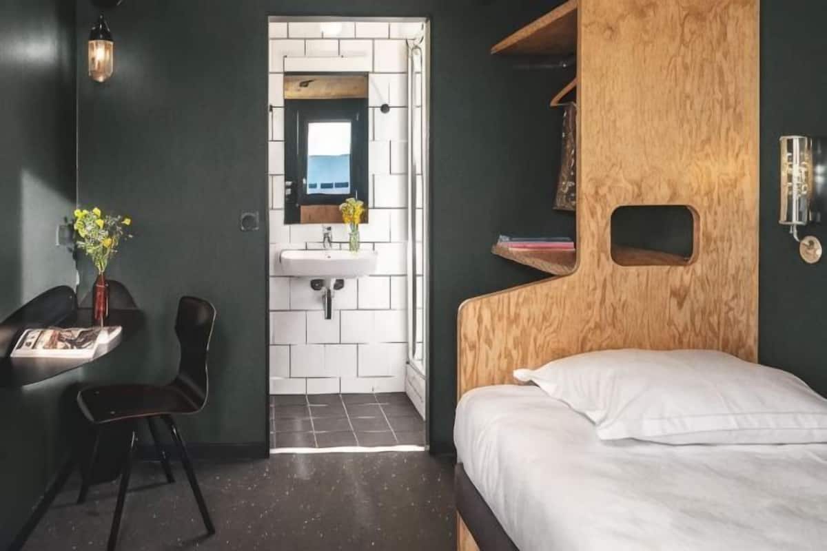 A bedroom with a wooden headboard, a white bed, a small desk with a chair, a wall-mounted shelf, and a bathroom with a white sink and mirror.