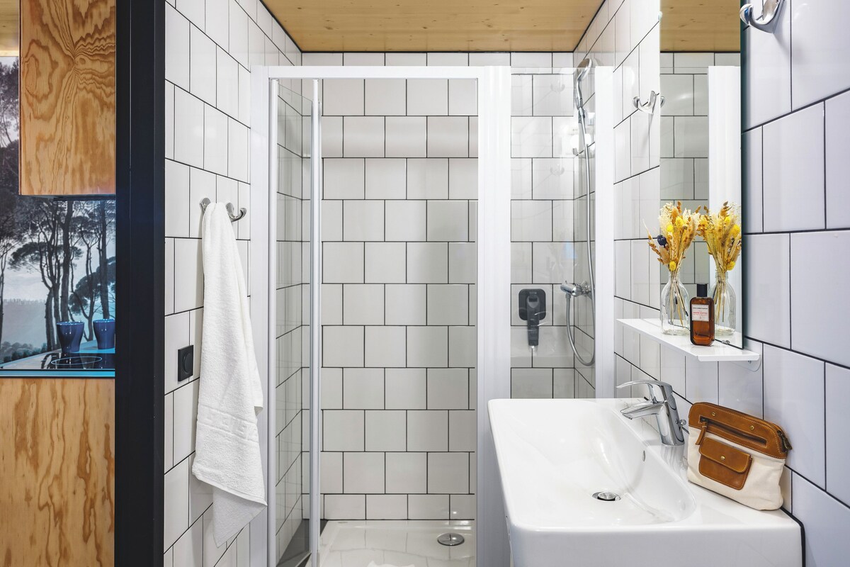 A modern bathroom with white tiled walls, a wooden ceiling, a white sink, a mirror, and a shower enclosure.