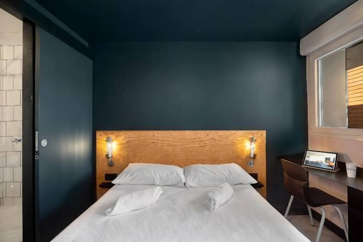 A hotel room with a wooden headboard, two white pillows, a neatly made bed, a bedside table with a lamp, and a window with blinds.