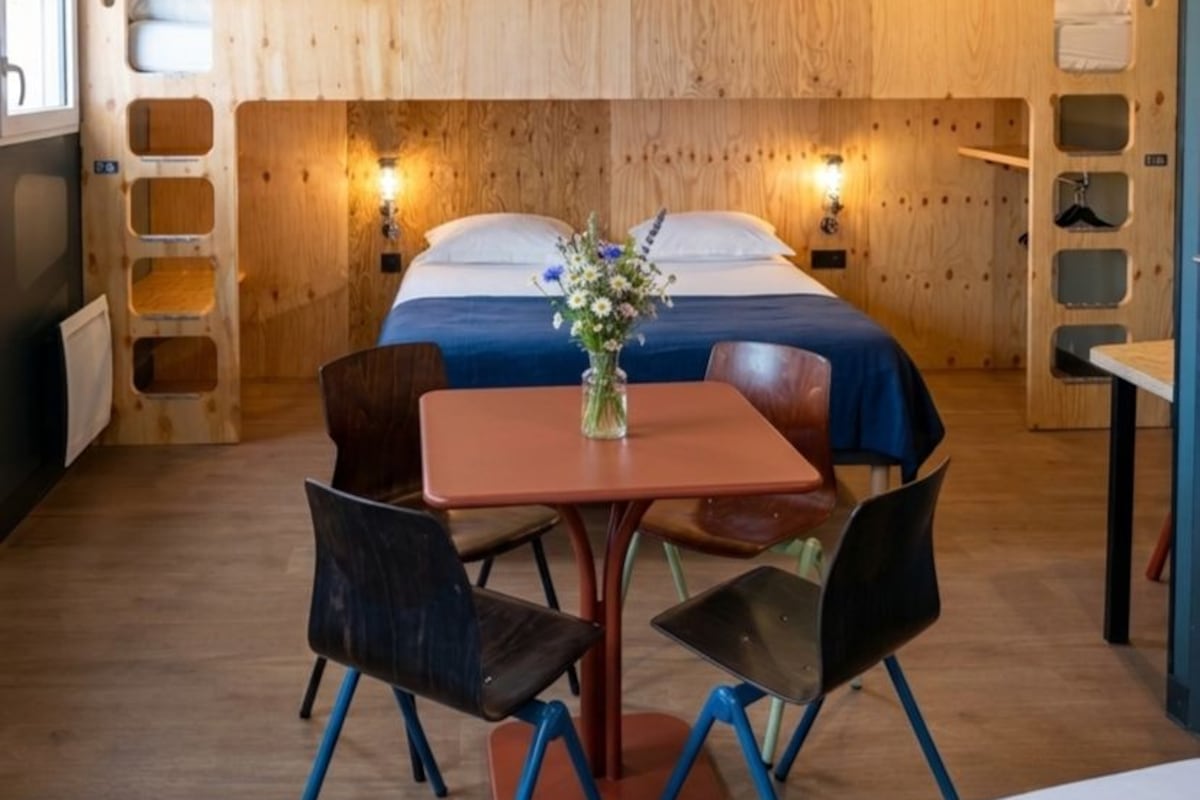 A room with a bunk bed, a table with chairs, and a vase with flowers.