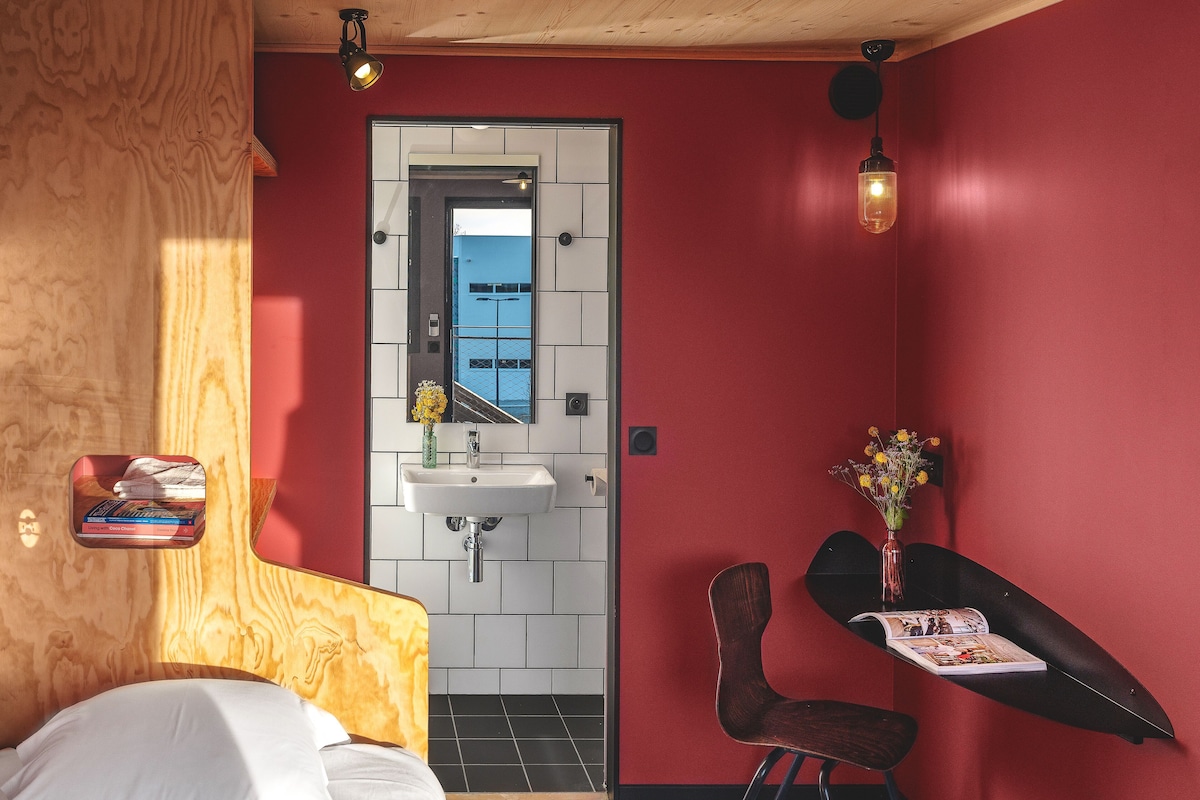 A hotel room with a bed, a small desk with a chair, a mirror, a sink, and a window.