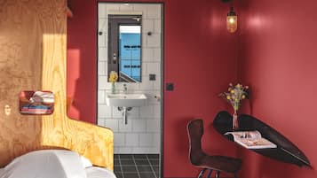 Een hotelkamer met een bed, een klein bureau met een stoel, een spiegel, een wastafel en een raam.