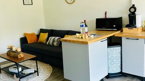 Ein modernes Wohnzimmer mit einem dunklen Sofa, einem Holztisch und einem kleinen Beistelltisch mit Kerzen. Ein Küchenbereich mit Mikrowelle und Wasserkocher ist sichtbar.