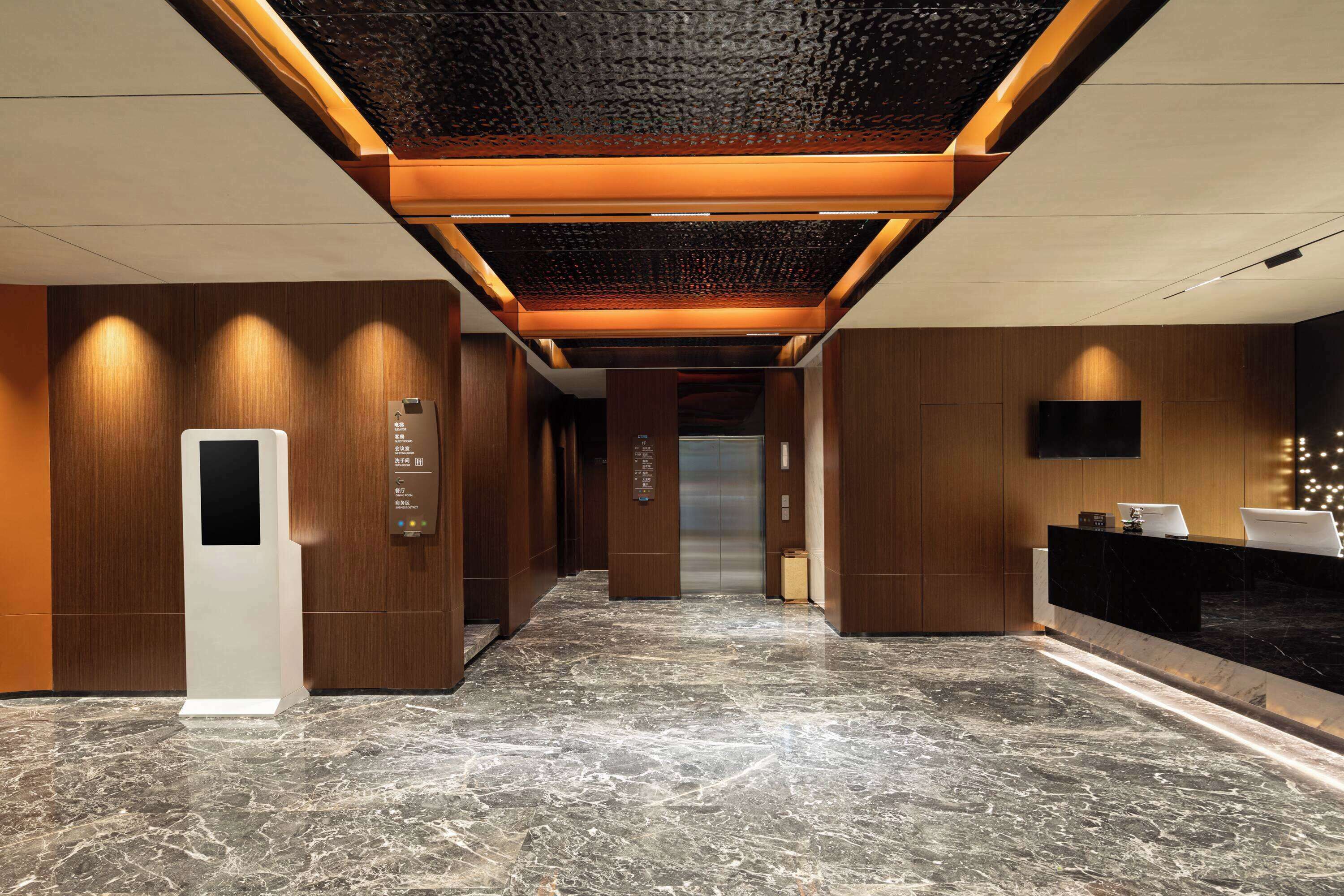 A modern hotel lobby with a marble floor, wooden paneling, a reception desk, and an elevator.