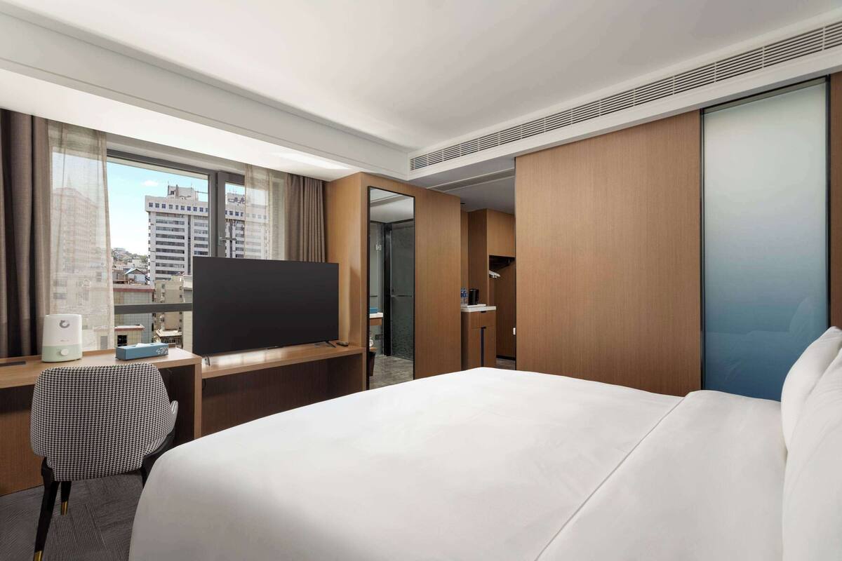 A hotel room with a large bed, a desk with a flat-screen TV, a chair, and a wardrobe.