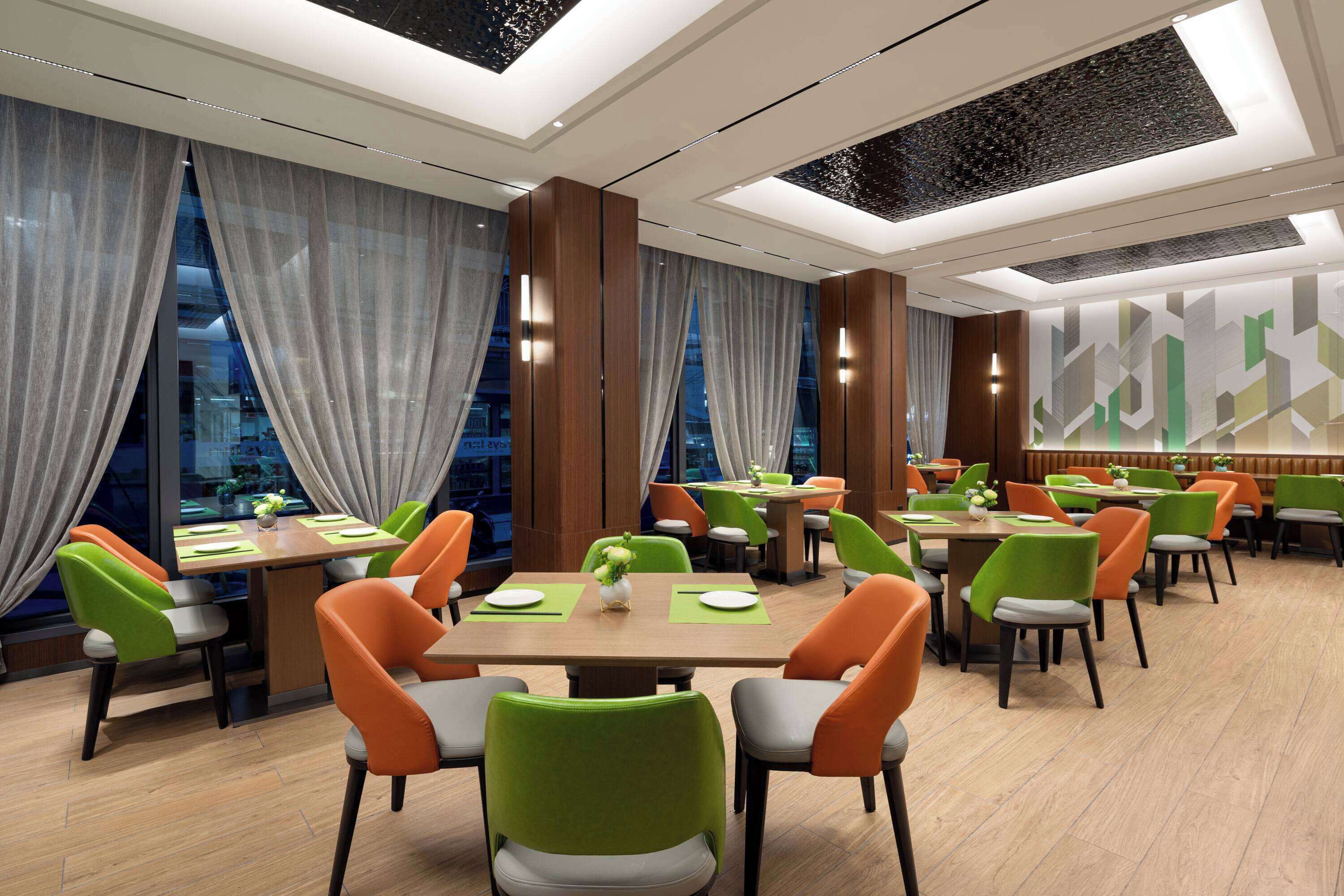A modern dining area with multiple tables and chairs, large windows with curtains, and a geometric wall design.