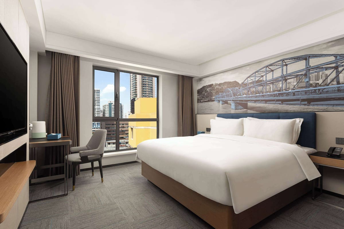 A hotel room with a large bed, a desk, a chair, a TV, and a window with a city view.