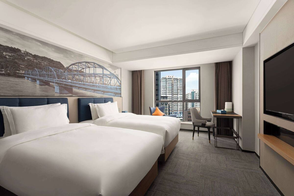 A hotel room with two beds, a desk, a chair, a TV, and a large window with a city view.