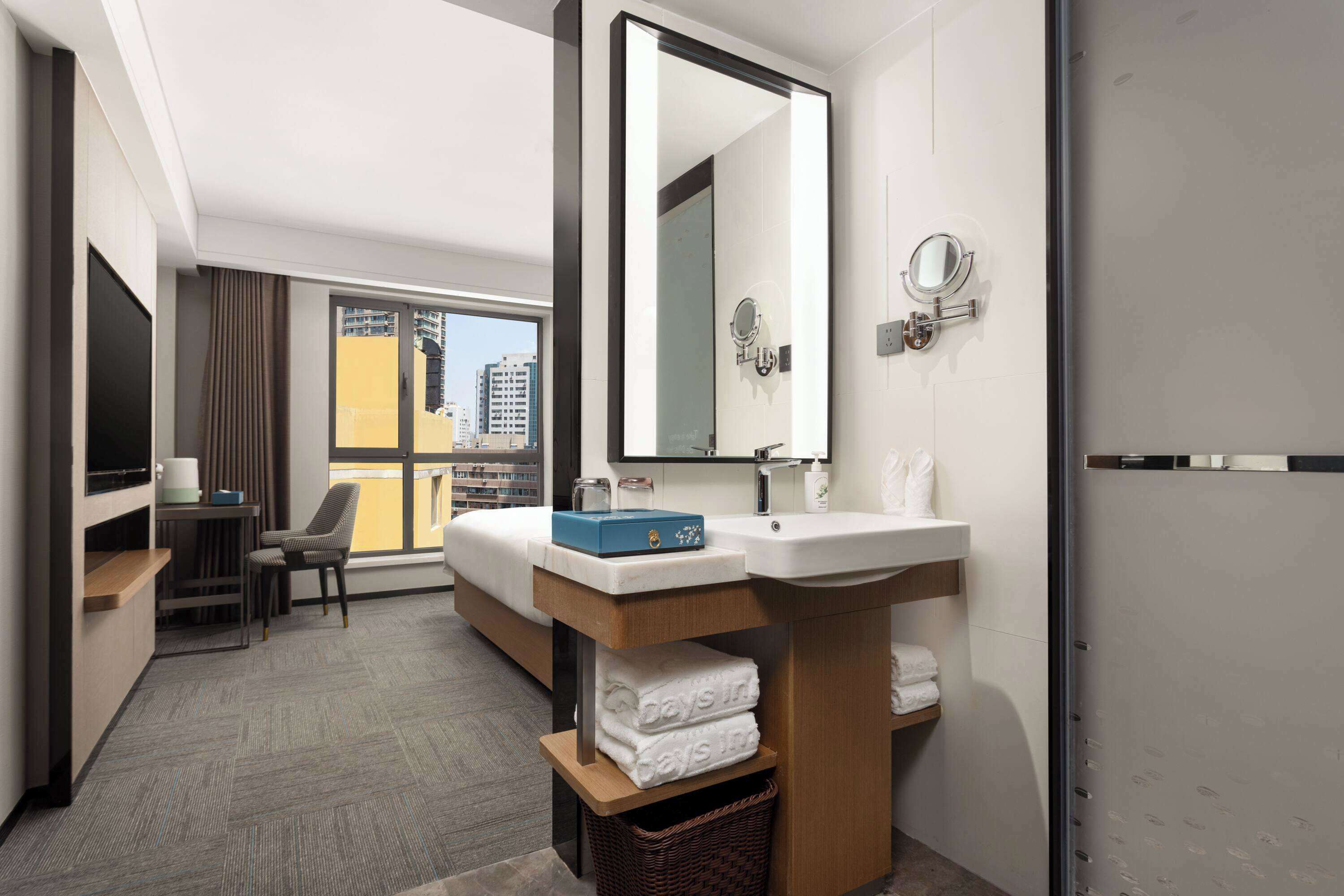 Una habitación de hotel moderna con una cama grande, un escritorio, una silla, un baño con lavamanos y espejo, y vistas de la ciudad a través de amplios ventanales.