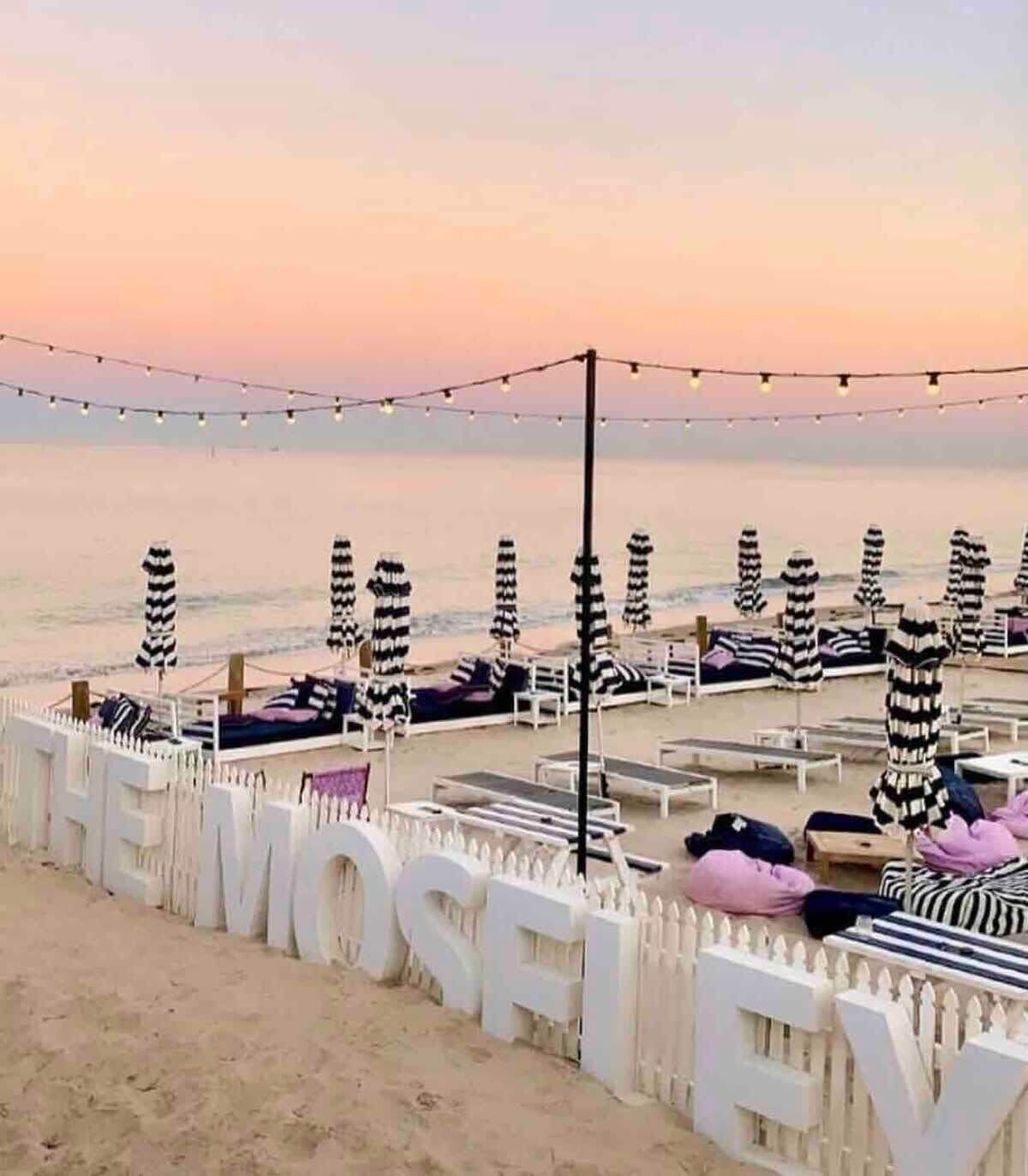 A beachside setting with striped umbrellas and lounge chairs, a white fence with the word "MOSFLEV" on it, and a string of lights in the background.