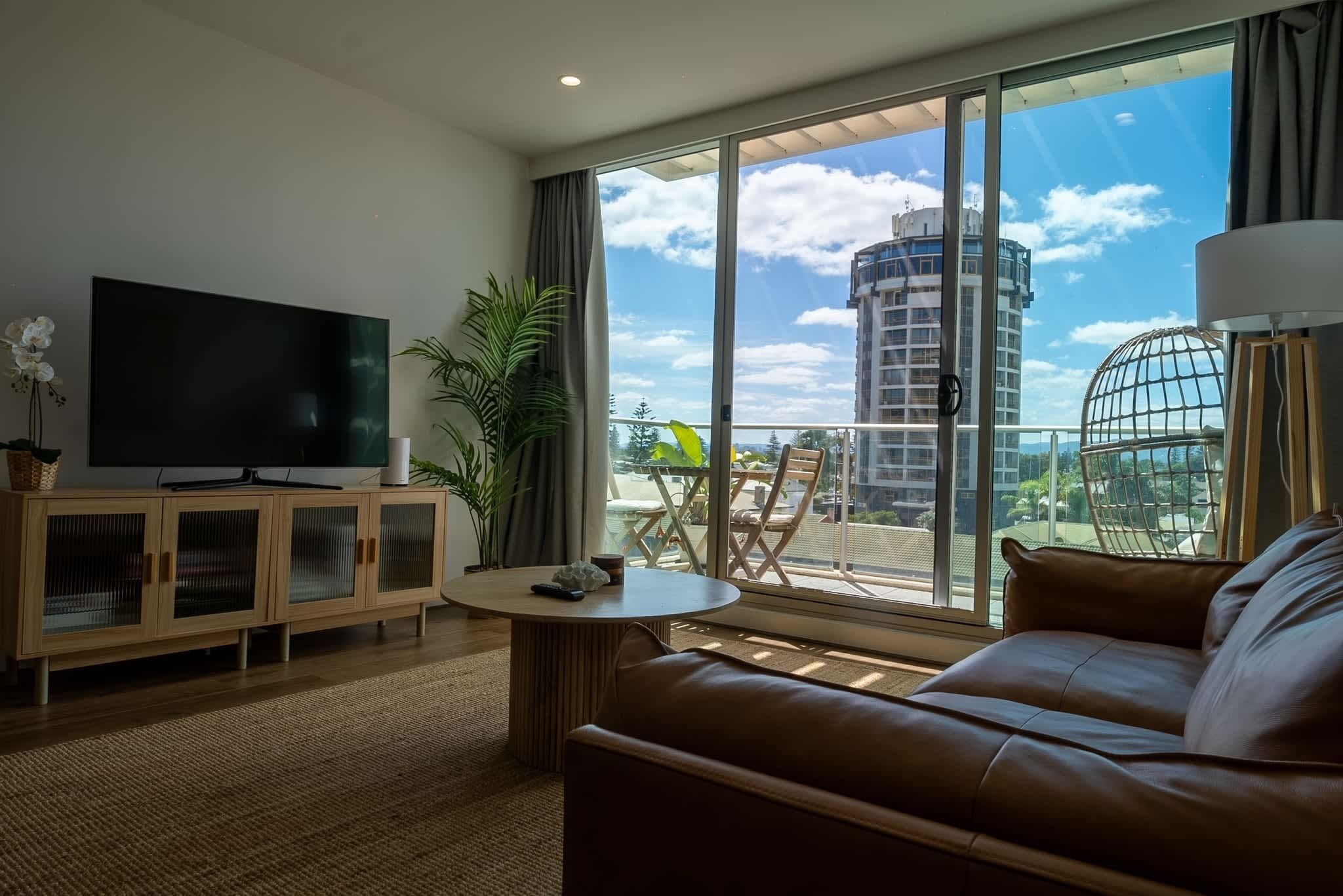 A modern living room with a large TV, a leather sofa, a round coffee table, and a balcony with a view of a cityscape.