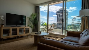 A modern living room with a large TV, a leather sofa, a round coffee table, and a balcony with a view of a cityscape. - Pool, Gym, Spa and Sauna, Free Parking, City Views (Glenelg)