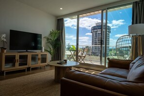 A modern living room with a large TV, a leather sofa, a round coffee table, and a balcony with a view of a cityscape. - Pool, Gym, Spa and Sauna, Free Parking, City Views (Glenelg)