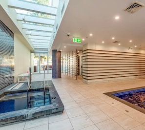 Et moderne poolområde indendørs med glastag, en lille pool og et loungeområde.